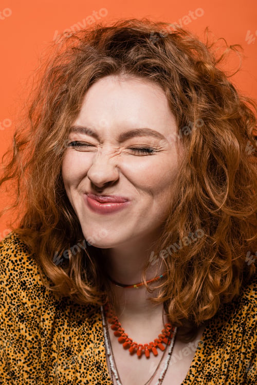 Preview: Pleased young red haired woman with natural makeup wearing necklaces and yellow blouse with print