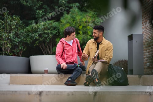 Preview: Happy young man with Down syndrome and mentoring friend sitting and talking outdoors