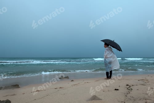 Preview: Young woman walking with an umbrella in front of ocean in rainy weather