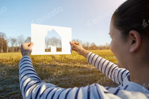 Preview: Woman standing in rural setting, holding out paper with cut-out of house