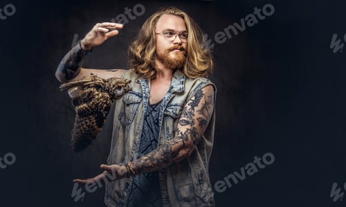 Preview: Portrait of a tattooed redhead hipster male holds a keeps the scarecrow of an owl in a studio