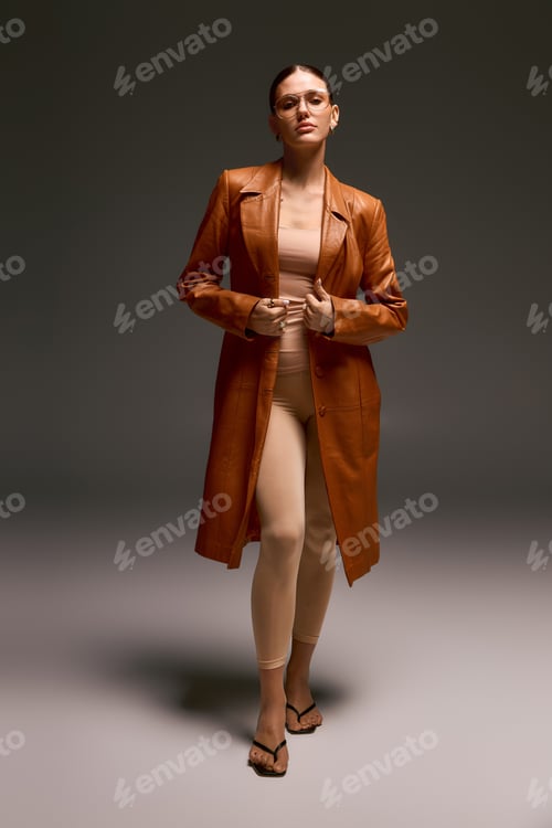 Preview: Stylish young woman in leather coat and glasses stands confidently in studio light