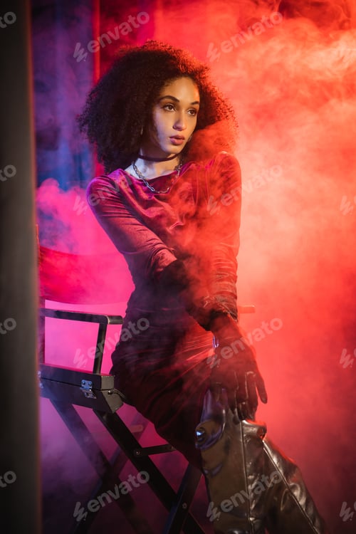 Preview: Trendy african american woman in velvet dress sitting on chair on red background with blue light