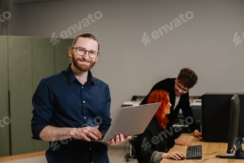 Preview: Smiling man holding laptop in modern office environment