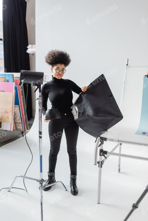 Preview: full length of smiling african american content maker in black clothes holding softbox near