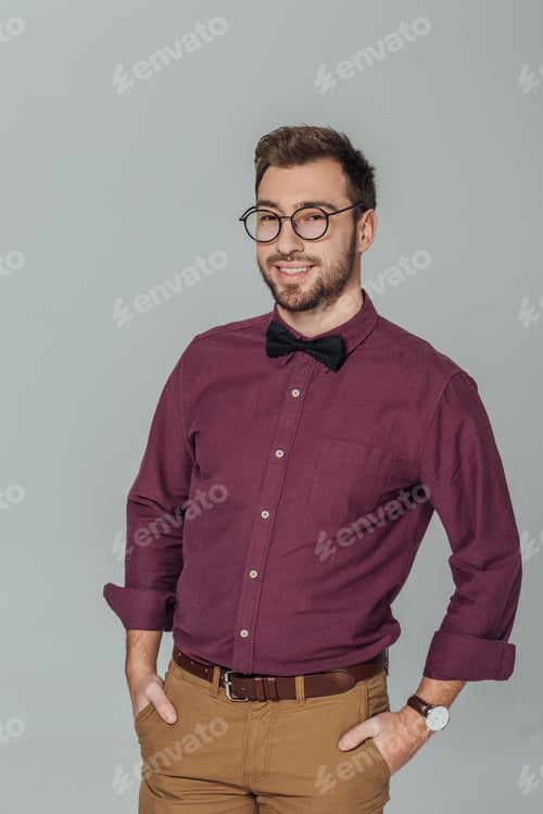 Preview: stylish young man in eyeglasses standing with hands in pockets and smiling at camera isolated on