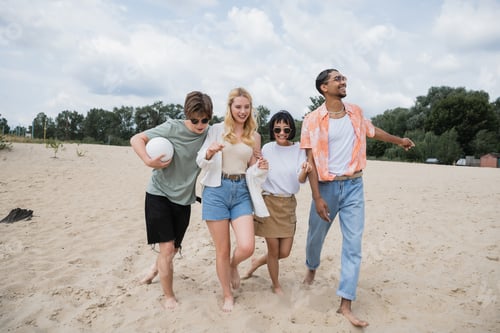 Preview: excited multicultural friends walking on sand beach with ball