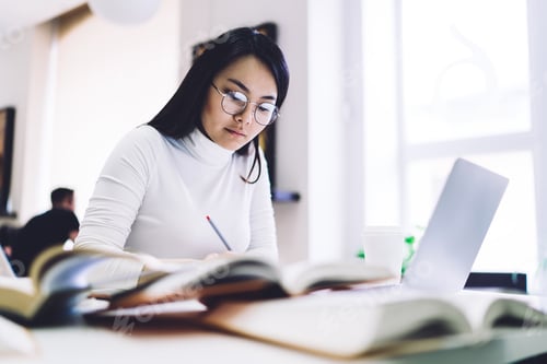 Preview: Pensive ethnic lady writing in notepad while sitting at desk with books and laptop in library