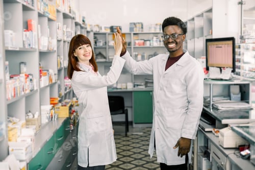 Preview: Female and male pharmacists in pharmacy. African man and Caucasian woman giving five, working at