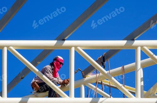 Preview: Construction worker is welding metal on top of building roof structure against blue sky