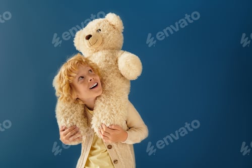 Preview: Blonde boy joyfully carries stuffed bear on his shoulders in a blue studio