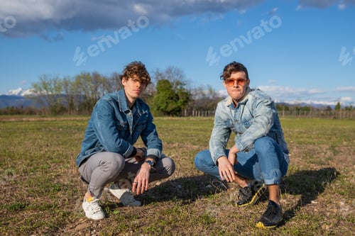 Preview: Two boys dressed in denim jackets pose on a meadow squatting together. Brotherhood and friendship