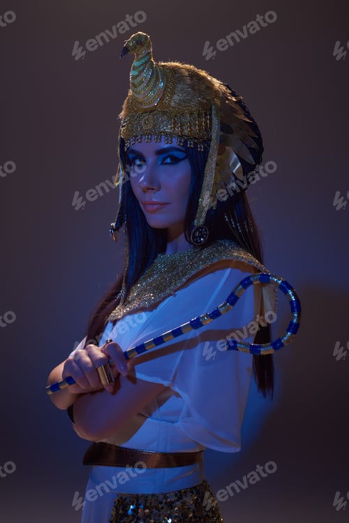 Preview: Portrait of woman in egyptian look holding crook and posing on brown background with blue light