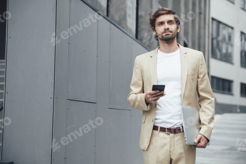 Preview: Thoughtful man in suit holding smartphone and laptop outside