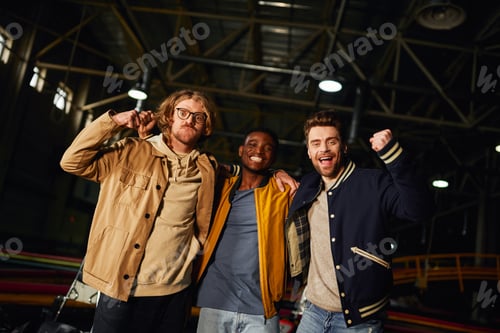 Preview: three interracial male friends smiling and standing near racing cars inside of indoor karting track
