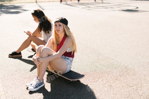Preview: Two adult female friends sitting on skateboards on basketball court