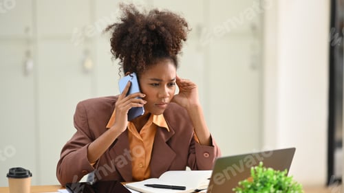 Preview: Serious African woman employee looking at laptop screen and talking on mobile phone.