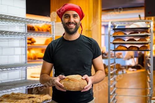 Preview: Corporate portrait of smiling bakery manager in the artisan bakery workshop, with a bread his hands