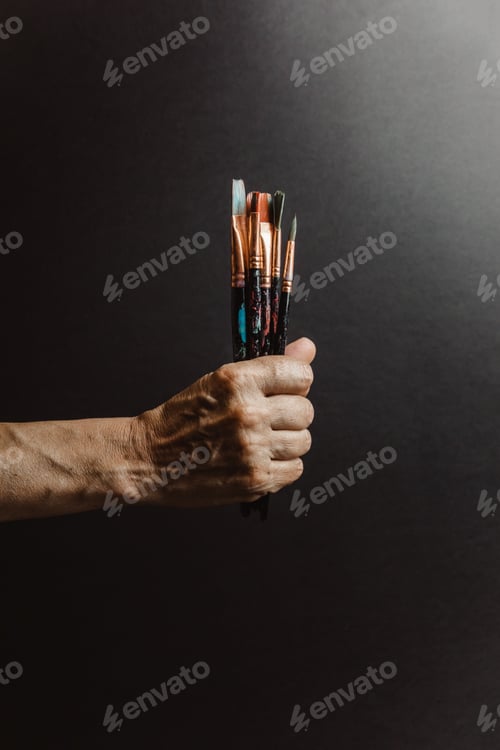 Preview: Hands of old female artist holding messy dirty paint brushes with different paints in art studio