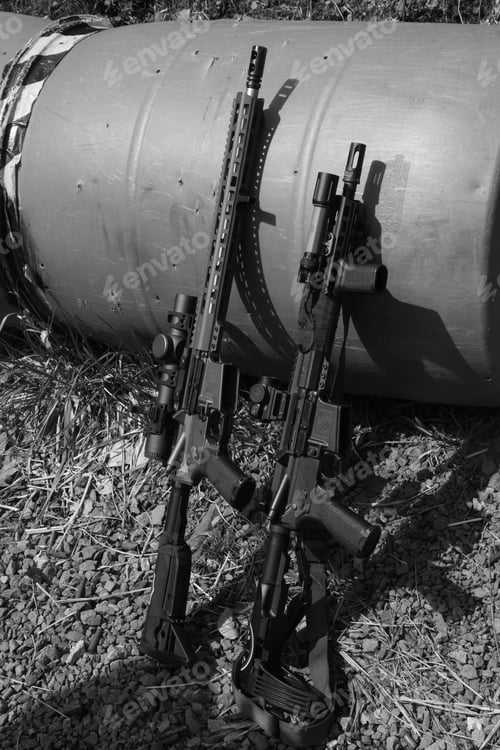 Preview: Grayscale shot of two handguns on the grass, positioned next to an empty barrel on the ground.