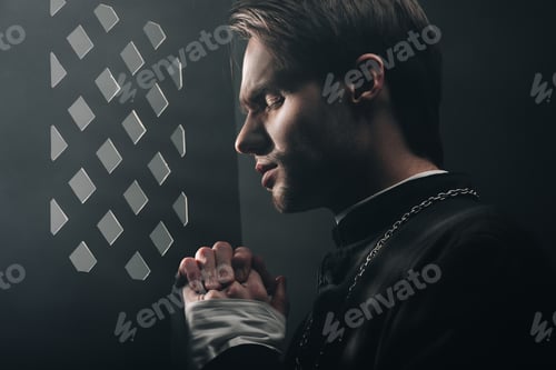 Preview: young thoughtful catholic priest praying with closed eyes in dark near confessional grille with rays