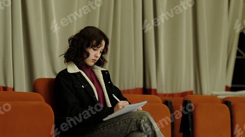 Preview: A young girl sits in a theater chair, focused on writing in a notebook. This image captures themes