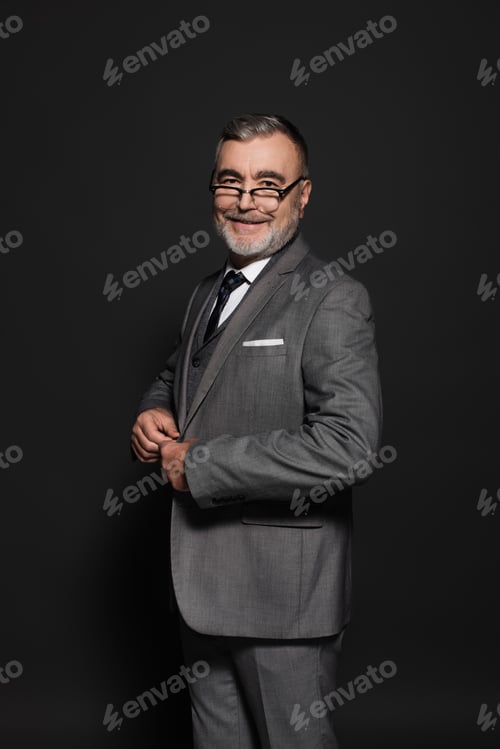 Preview: cheerful senior man buttoning blazer while looking at camera on dark grey