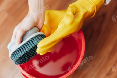 Preview: Person in yellow rubber gloves cleaning red bucket with brush on wooden floor next to bucket of