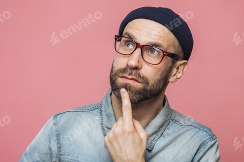 Preview: Close up shot of pleasant looking bearded male with thoughtful expression, keeps finger on chin