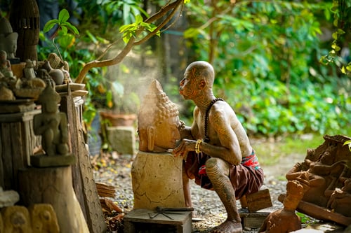 Craftsman shaping a stone Buddha statue in a lush garden in Thailand during daylight