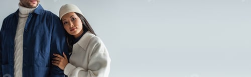 Preview: pretty asian woman in white jacket and beanie leaning on man isolated on grey, banner