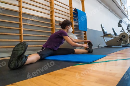Preview: Young caucasian girl with face mask doing stretching after finishing the exercise in a gym