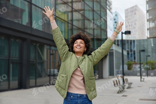 Vista previa: Horizontal shot of happy carefree woman with curly hair keeps arms raised up enjoys life poses durin