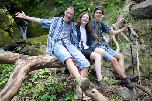 Preview: Group of friends with backpacks and sticks sitting on a fallen tree, Take a break during the hike