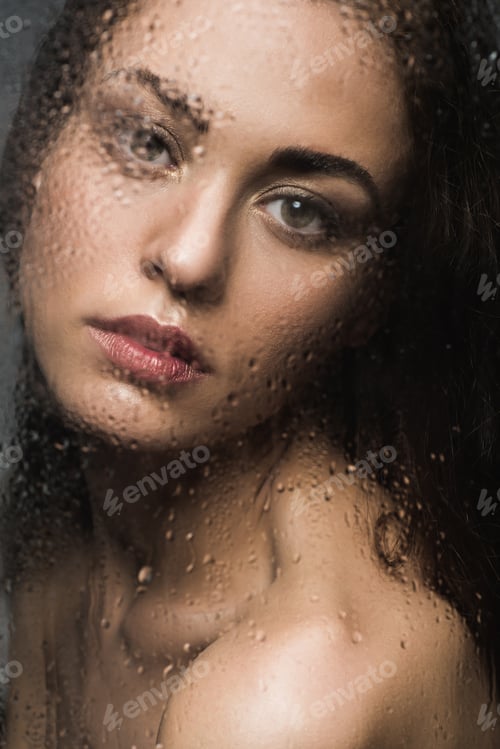 Preview: attractive girl behind wet glass looking at camera