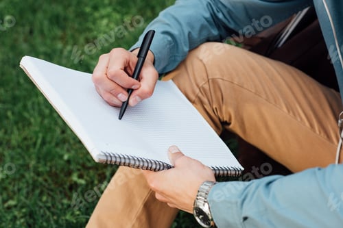 Preview: cropped shot of young man writing in blank notebook while sitting on grass