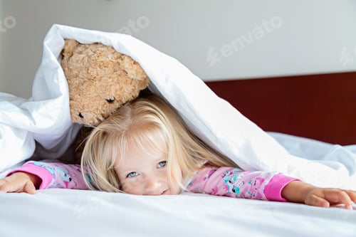 Preview: Cute little Caucasian girl with blonde hair wearing pajamas and lying in bed with stuffed teddy bear