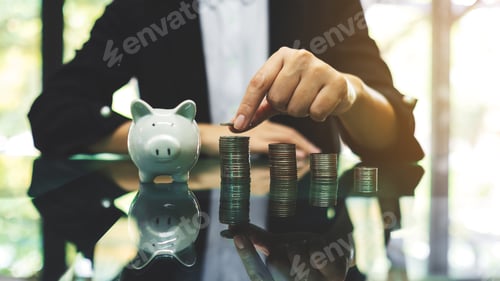 Preview: Businesswoman stacking coins with a white piggy bank on the table for saving money