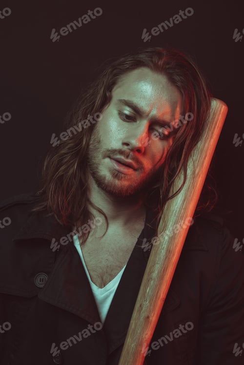 Preview: Serious tired young man with long hair holding baseball bat and looking down