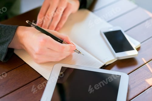 Preview: Individual writing notes with a pen on a notebook, surrounded by digital devices on a wooden table