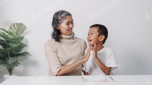 Preview: Asian grandson and grandmother people sitting together at desk doing homework at home.