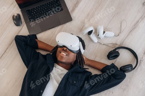 Preview: Computer nearby. Top view of man that is lying down on floor in virtual reality headset