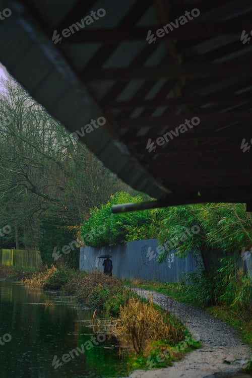 Preview: Vertical from under a shelter, man with an umbrella walking on a pathway near a river under the rain