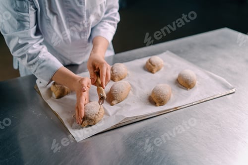 Preview: woman baker cuts patterns raw bread rolls with a professional baker's knife before baking