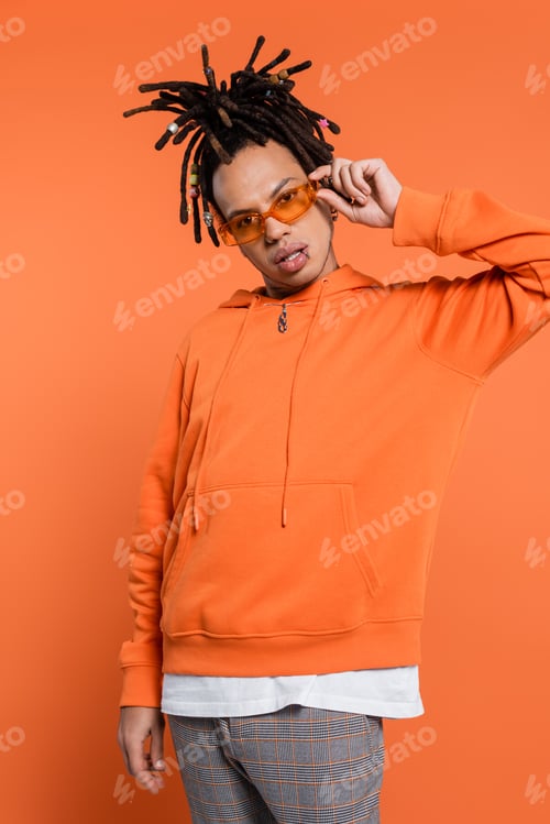 Preview: pierced multiracial man with dreadlocks adjusting trendy sunglasses on coral background