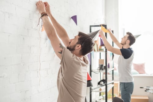 Preview: side view of two young men in party hats hanging up string with party garlands on brick wall