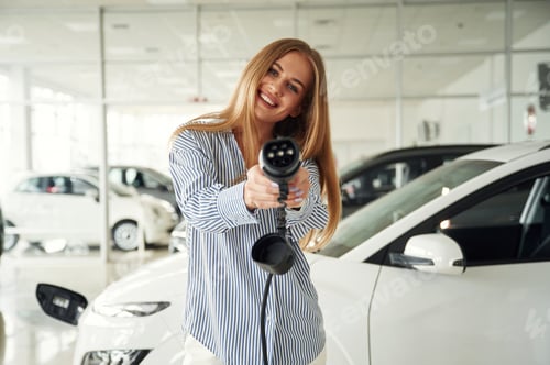 Preview: Smiling and holding charging device for electric car. Beautiful woman is indoors