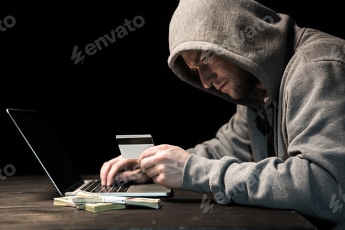 Preview: Side view of young man in hoodie holding credit card and using laptop