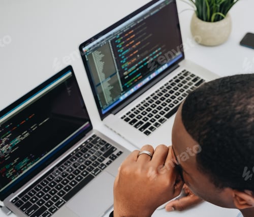 Preview: african american man sitting at two laptop computers at white desk looking at software code