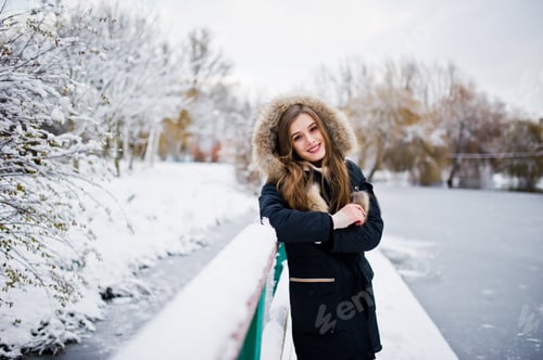 Preview: Beautiful brunette girl in winter warm clothing. Model on winter jacket against frozen lake at park.